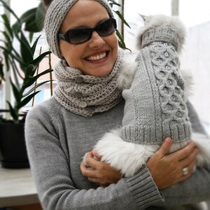 May include: A woman wearing a gray knitted headband and scarf holds a white fluffy dog wearing a gray knitted sweater. The dog is wearing a gray knitted sweater with a cable knit pattern.