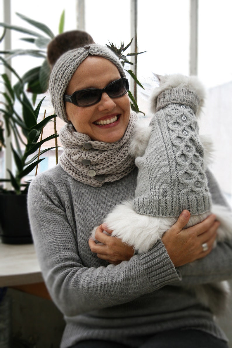 May include: A woman wearing a gray knitted headband and scarf holds a white fluffy dog wearing a gray knitted sweater. The dog is wearing a gray knitted sweater with a cable knit pattern.