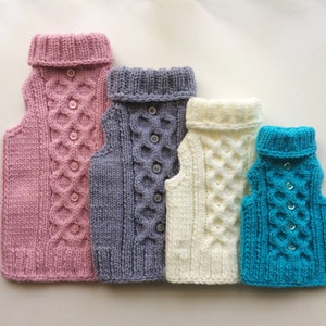 May include: Four knitted dog sweaters in different colors: pink, gray, white, and turquoise. Each sweater has a turtleneck and a cable knit pattern with buttons down the front.