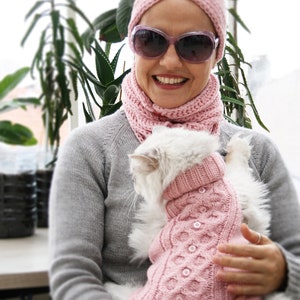 May include: A woman wearing a pink headband and a pink scarf is holding a white cat wearing a pink knitted sweater. The cat is sitting on the woman's lap.