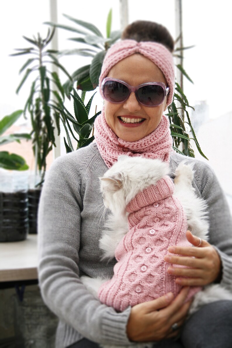 May include: A woman wearing a pink headband and a pink scarf is holding a white cat wearing a pink knitted sweater. The cat is sitting on the woman's lap.