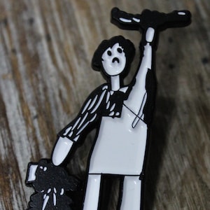 Puede incluir: Pin de esmalte blanco y negro de una persona sosteniendo un palo con un perro a su lado. La persona lleva una camisa a rayas y un delantal blanco. El perro es negro.