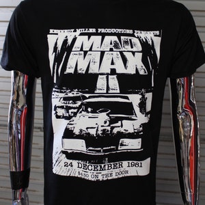 May include: Black t-shirt featuring a white graphic of the movie "Mad Max II". The graphic includes the movie title, car illustrations, and the date "24 December 1981". The shirt is displayed on a mannequin.