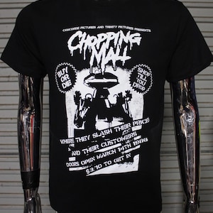 May include: Black t-shirt featuring a white graphic for "Chopping Mall." The design includes the film's title in a dripping font, a robot illustration, and text that reads "Buy or Die" and "Shop Till You Drop."