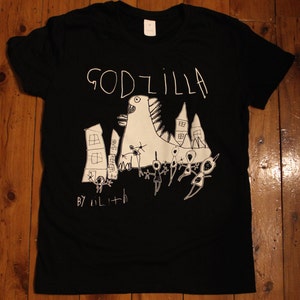 May include: Black t-shirt featuring a white drawing of Godzilla destroying a city. The drawing is in a childlike style and says "GODZILLA" at the top and "BY LILITH" at the bottom.