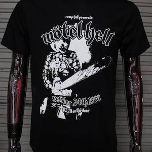 以下が含まれることがあります： 白のグラフィックがプリントされた黒いTシャツ。グラフィックには、チェックシャツを着てチェーンソーを持った人が描かれている。グラフィックの上には「Motel Hell」という文字が、下には「October 24th 1980 2.00 ドル at the door」という文字が書かれている。