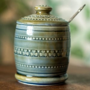 May include: A ceramic jar with a lid and a small spoon. The jar is cylindrical with a light blue and green colour scheme. It features horizontal bands with dotted and clover patterns. The jar sits on a small base.