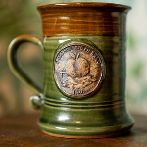 May include: Green ceramic mug with a brown rim and handle. The mug features a circular emblem with an apple and the text "Goodhill Cider Barn". The mug is on a wooden surface.
