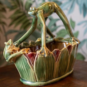 May include: A ceramic basket featuring a glossy green finish with sculpted frog figures. One frog forms the arched handle, while another smaller frog is perched on the rim. The exterior is decorated with vertical leaf-like patterns in shades of green and brown.
