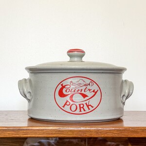 May include: A white ceramic casserole dish with red lettering that reads "Country Pork". The dish has two handles and a lid.