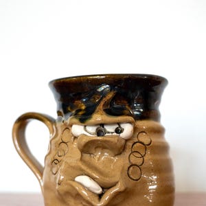 May include: A handmade ceramic mug with a face design. The mug is mainly tan with a dark brown rim and handle. The face has white eyes, a large nose, and a protruding white tooth. Decorative circles are on the side.