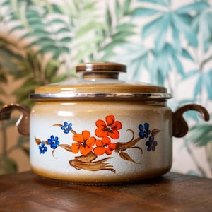 May include: A vintage enamel cooking pot with a glass lid. The pot is cream-coloured with a brown rim and wooden handles. It features a floral design with orange and blue flowers. The pot is on a wooden surface.