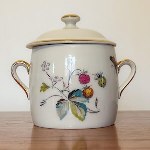 May include: A white ceramic sugar bowl with a lid and gold handles. The bowl is decorated with a floral pattern featuring pink, yellow, and green strawberries.