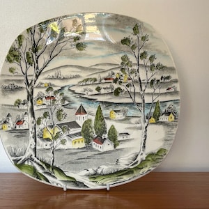 May include: A vintage ceramic plate featuring a scenic design of a small town with houses, trees, and a river. The plate is white with black, green, yellow, and red accents.