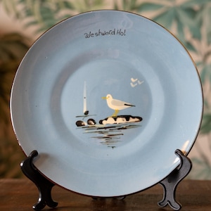 May include: A light blue ceramic plate featuring a hand-painted scene of a white and yellow seagull perched on a dark rock. The text "Westward Ho!" is written in black script near the top rim, accompanied by a small sailboat and white birds.