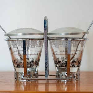 May include: A vintage salt and pepper set with a metal frame and lids. The clear glass shakers feature a geometric black and silver design. Each shaker has a small spoon. The set sits on a wooden surface.