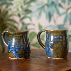May include: Two ceramic mugs featuring a reactive glaze finish with deep olive green and vibrant cobalt blue tones. Each mug has a rounded body, a sturdy side handle, and a smooth, glossy surface texture with visible colour blending and drips.