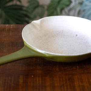 May include: A round frying pan with a smooth, olive green exterior and a matte, off-white interior surface. The pan features a long, integrated handle with a small circular hole at the end and a small pouring spout on the rim of the cooking vessel.
