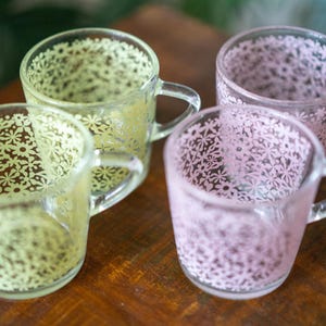 May include: Four clear glass mugs with handles, each decorated with a delicate floral pattern. Two mugs have a pale yellow tint, while the other two are a soft pink. The mugs are arranged on a wooden surface.