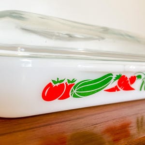 May include: White glass baking dish with a clear glass lid. The dish has a red and green vegetable design on the front. The design includes tomatoes, courgettes, and carrots.