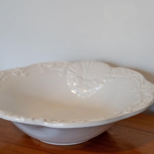 May include: A white ceramic serving bowl with a scalloped edge and embossed floral and shell designs. The bowl sits on a wooden surface, with a plain white wall in the background.