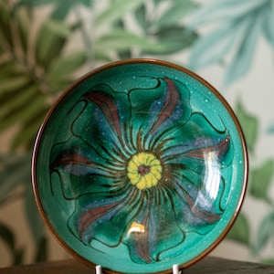 May include: A ceramic bowl with a floral design. The bowl has a teal background with a large flower in shades of green, brown, and yellow. The rim is a dark brown colour.