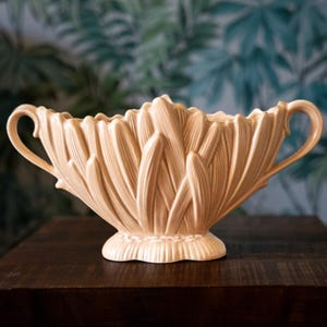 May include: A pale peach-coloured ceramic bowl featuring a textured, leaf-like exterior pattern. The vessel has a scalloped rim, a pedestal base, and two curved handles extending from the sides, creating a symmetrical, elongated silhouette.