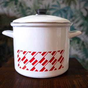 May include: A white enamel stockpot with a matching lid featuring a black knob. The pot has two side handles and a decorative band around the centre consisting of a repeating red and white geometric pattern of slanted rectangles and lines.