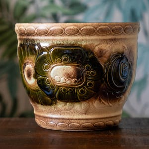 May include: A ceramic planter featuring a glossy glaze in shades of olive green and tan. The exterior is decorated with a repeating embossed floral pattern and a textured chain-link border along the top and bottom rims of the cylindrical vessel.