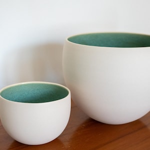 May include: Two white ceramic bowls with a teal interior. The bowls are different sizes and have a rounded shape. The bowls are on a wooden surface.