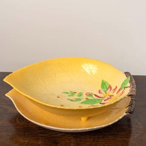 May include: A yellow leaf-shaped serving dish with a matching base. The dish features a floral design with pink and green accents. The dish is made of ceramic and has a brown handle.