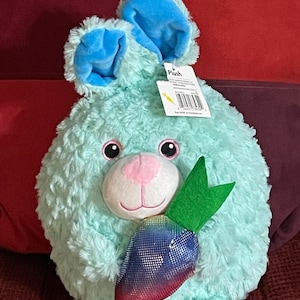 Puede incluir: Un conejito de peluche azul sosteniendo una zanahoria morada brillante con hojas verdes. El conejito tiene orejas azules y una nariz rosa. La etiqueta en el conejito dice "Plush".