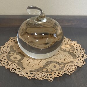 May include: A clear glass apple shaped paperweight with a curved stem. The paperweight is sitting on a beige lace doily.