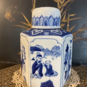 Puede incluir: Un tarro de cerámica blanco y azul con tapa. El tarro tiene forma octogonal y está decorado con ilustraciones azules de figuras y paisajes. El tarro está sobre un tapete de encaje blanco.