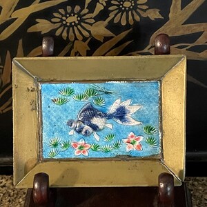 Puede incluir: Obra de arte rectangular con marco de latón, con un fondo azul y un diseño de pez dorado. El pez es blanco y azul, rodeado de plantas acuáticas verdes y flores rosas. La obra de arte se exhibe sobre un soporte de madera oscura.