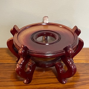 Puede incluir: Un soporte de exhibición de madera con una parte superior circular y patas curvas ornamentadas. El soporte está teñido en un rico color marrón rojizo, con un tono de madera más claro en la superficie superior. Diseñado para sostener un objeto decorativo.