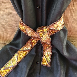 May include: A black button-down shirt with a decorative sequined belt. The belt is a ribbon of gold and bronze sequins, with a geometric pattern. The shirt is made of a shiny, flowing fabric.