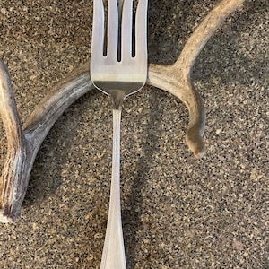 Op de afbeelding: Een zilveren serveervork met vier tanden, elk met een gebogen, puntige punt. De steel is lang en slank, met een decoratief bloemmotief aan de basis. De vork staat tegen een gespikkelde, grijze achtergrond.