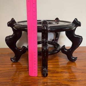 May include: A dark brown, wooden decorative stand with a round top and three curved legs. The stand is approximately 11 inches tall. A pink ruler is placed next to the stand for measurement.