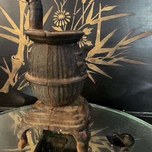 Puede incluir: Una pequeña estufa antigua de hierro fundido con una chimenea curva y una pequeña olla separada. La estufa es de color marrón oscuro con una superficie texturizada y se asienta sobre una base decorativa. El fondo presenta una pantalla negra con diseños florales y de bambú dorados.