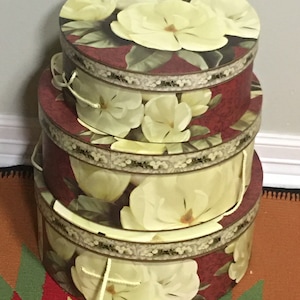 May include: Three round decorative boxes stacked on top of each other. The boxes are burgundy with a floral design of cream-coloured magnolia blossoms and green leaves. Each box has a decorative border and a rope handle.