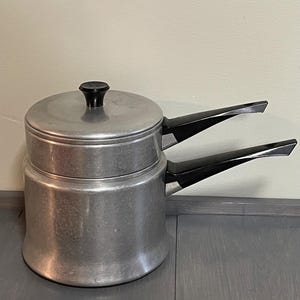 Puede incluir: Una olla doble de plata con dos asas negras. La olla superior tiene una tapa con un pomo negro.