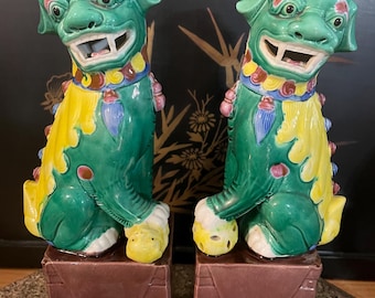 May include: Pair of green ceramic Foo Dogs with yellow and red accents, perched on brown square pedestals. The Foo Dogs have smiling expressions and hold yellow balls. Decorative Asian-inspired figurines.