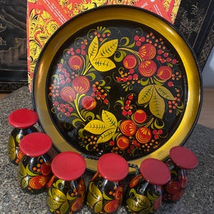 Puede incluir: Una bandeja de servicio redonda de color negro con borde dorado y un patrón floral pintado a mano con bayas rojas y hojas amarillas, acompañada de seis tazas pequeñas a juego con bases rojas y cuerpos negros decorados con motivos florales.