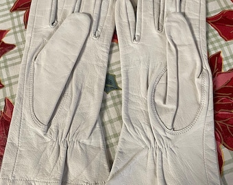 May include: A pair of off-white leather gloves. The gloves are laid out flat, showcasing the details of the stitching and the supple texture of the leather. The gloves have a classic design, with a gathered wrist and a smooth finish.