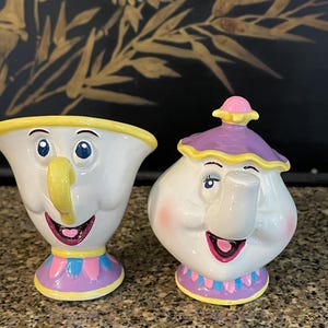 May include: Two ceramic figurines of characters from Beauty and the Beast. One is a white teacup with a yellow handle and a smiling face. The other is a white teapot with a purple lid and a smiling face. Both have pink, blue, and yellow accents.