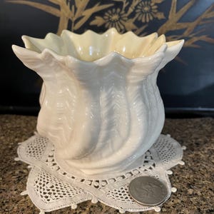 Può includere: Un vaso di ceramica bianca con bordo a balze, appoggiato su una tovaglietta di pizzo bianco. Il vaso presenta un motivo a spirale e strutturato. Una moneta d'argento è accanto alla tovaglietta. Lo sfondo è scuro con dettagli floreali dorati.