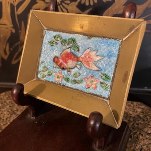 Puede incluir: Una pieza decorativa rectangular con marco dorado que presenta un diseño de pez dorado. La obra de arte muestra un pez dorado rojo y blanco nadando en un estanque azul con plantas verdes y flores rosas. La pieza se exhibe en un soporte de madera marrón oscuro.