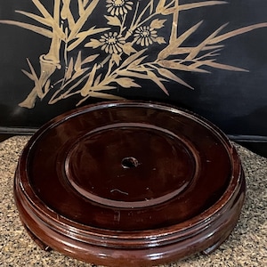 Op de afbeelding: Een ronde, donkerbruine houten displaystandaard met een glanzende afwerking. De standaard heeft een verhoogd midden en een decoratieve, gelaagde rand. De achtergrond toont een zwart paneel met gouden bloemen- en bamboe-ontwerpen.