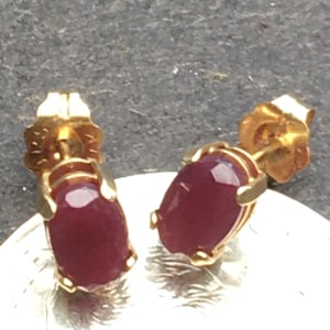 May include: A pair of gold stud earrings, each featuring a deep red oval gemstone. The gemstones are set in a four-prong gold setting. The earrings are displayed on a silver surface.